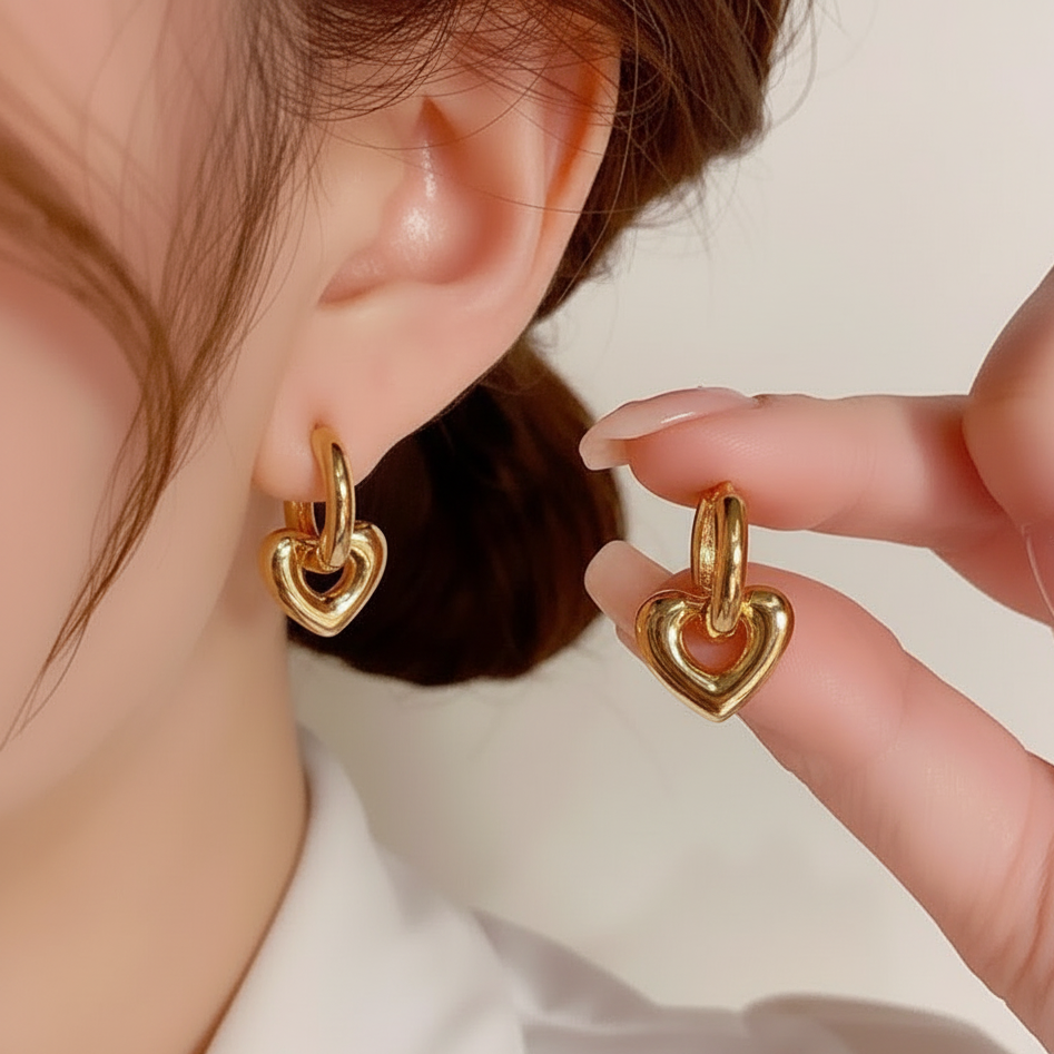 A close-up lifestyle shot of a person wearing a gold heart-shaped hoop earring, with their other hand holding up a matching gold heart hoop earring for display.